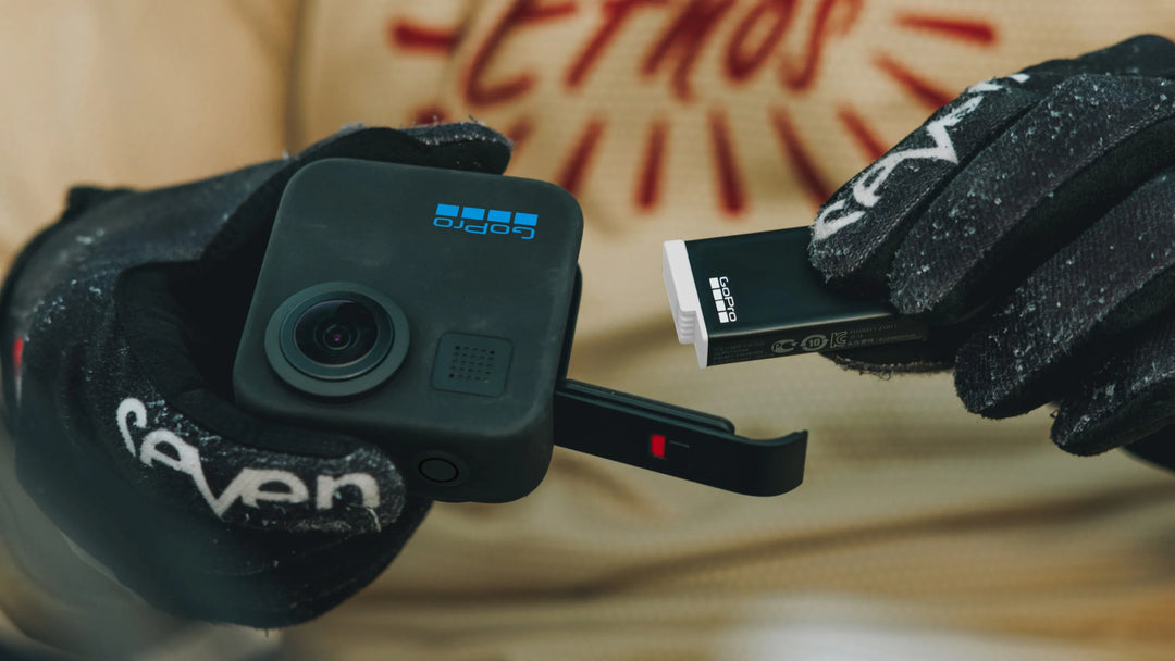 Person holding a small camera device with a USB stick, wearing gloves, against a blurred background.