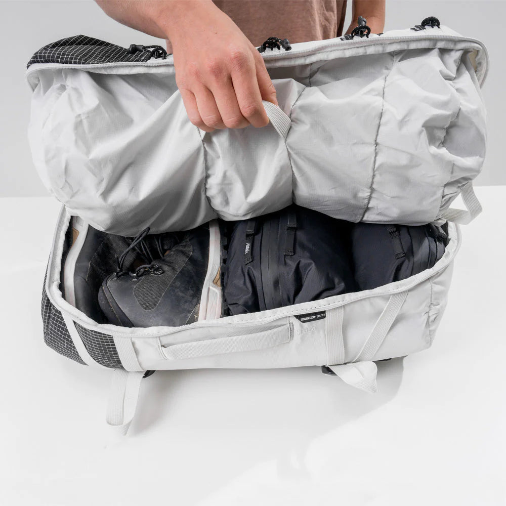 An image of an open white backpack on a white surface, with a hand pulling back the top flap. Inside the backpack, there are a pair of black shoes with laces and several black packed items, neatly arranged.