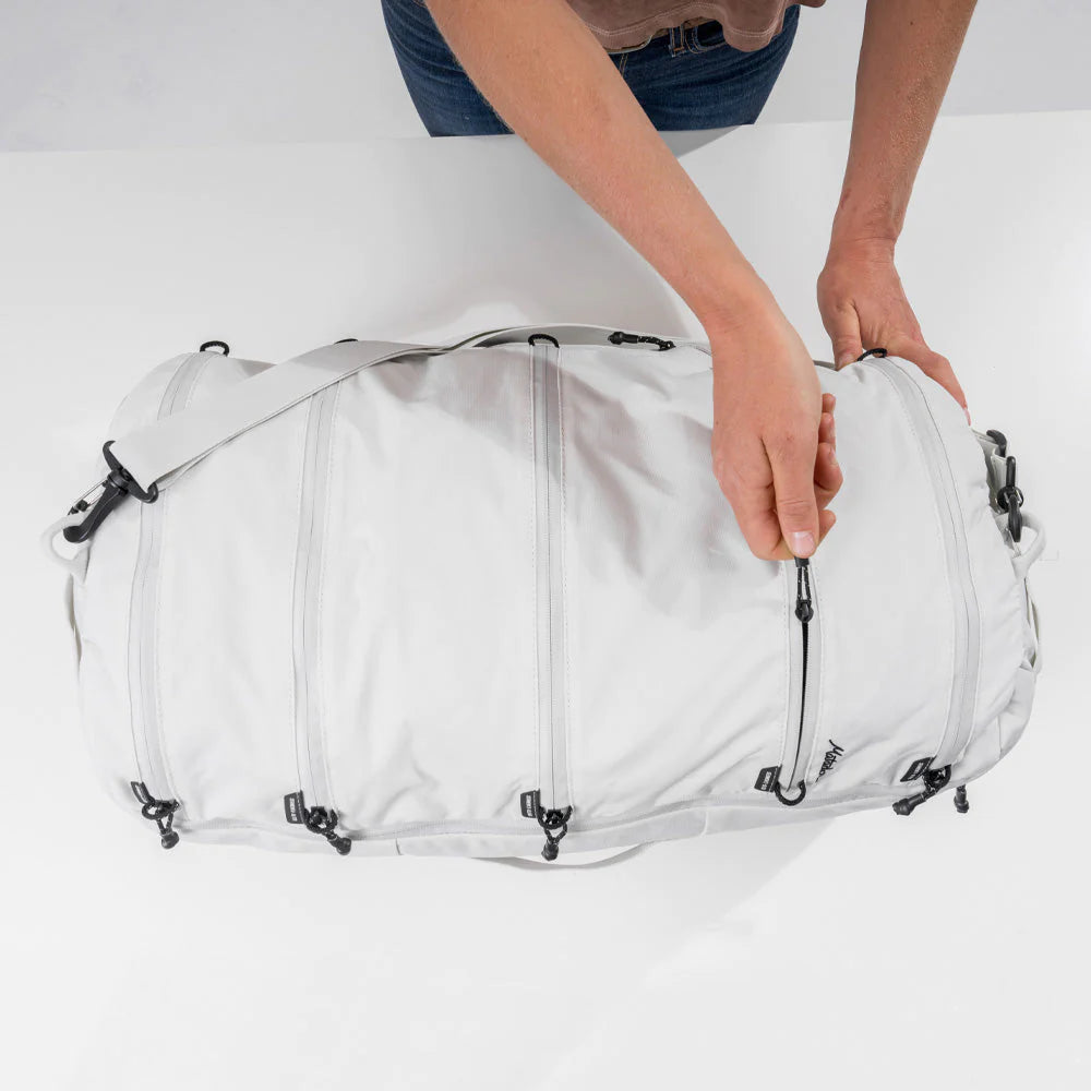 An image of a person opening a white duffel bag on a white surface, with both hands adjusting the top flap. The bag features multiple compartments with black straps and buckles, and the brand name "Matador" is visible on the side.