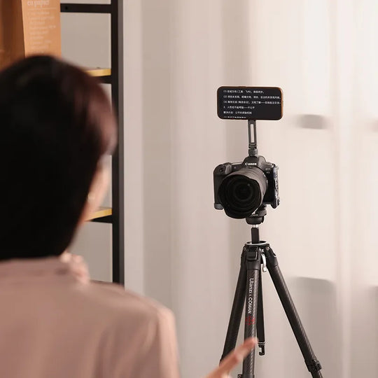 Person using a camera on a tripod with a microphone attached, in a room setting.
