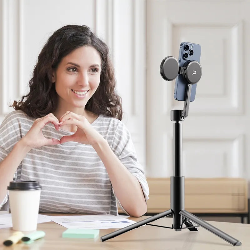 Woman sitting at a table with a smartphone on a stand, making a heart shape with her hands.