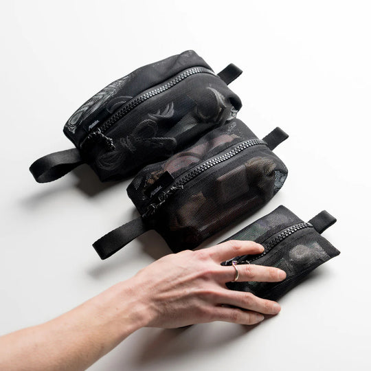 Set of black mesh toiletry bags with a hand reaching for one on a light gray background