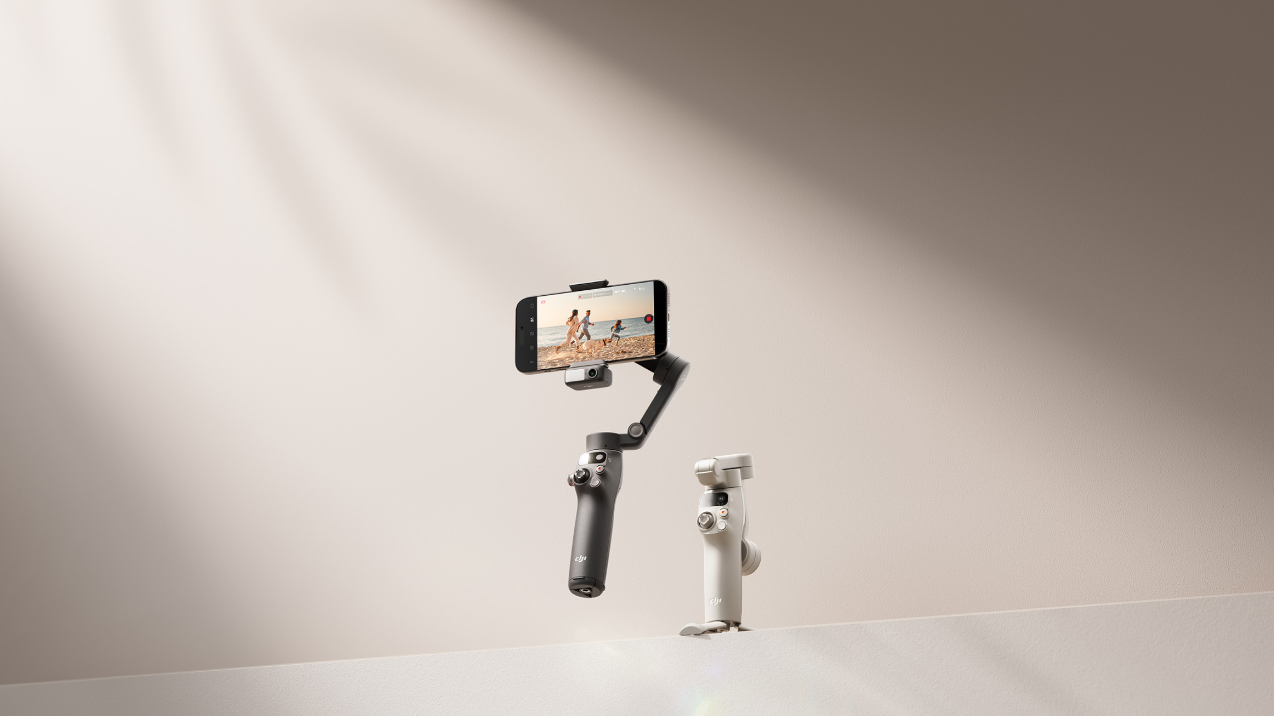 A black DJI Osmo Mobile 7 gimbal with a smartphone attached is positioned slightly above and to the left of a white DJI Osmo Mobile 7 gimbal, both floating on a light brown background. The smartphone screen displays two people walking on a beach. The text 