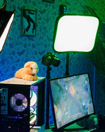 A vibrant gaming or streaming setup with a glowing computer tower, a camera on a tripod, and two softbox lights. The room is illuminated with green and blue light, and a monitor in the foreground displays a video game. A small plush toy rests on top of the computer tower.