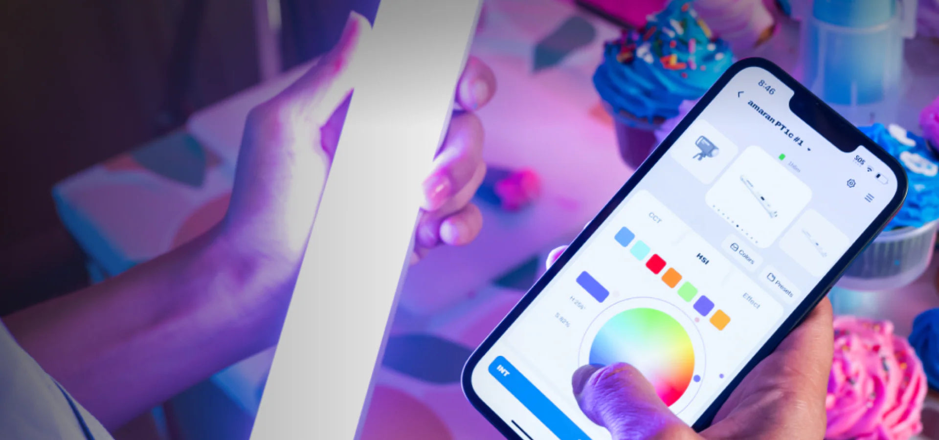 A close-up shot of a person's hands holding a smartphone, which displays the Amaran PT 1c lighting control app with a color wheel and various sliders. In the background, another hand holds a white light stick, with cupcakes visible on a table.