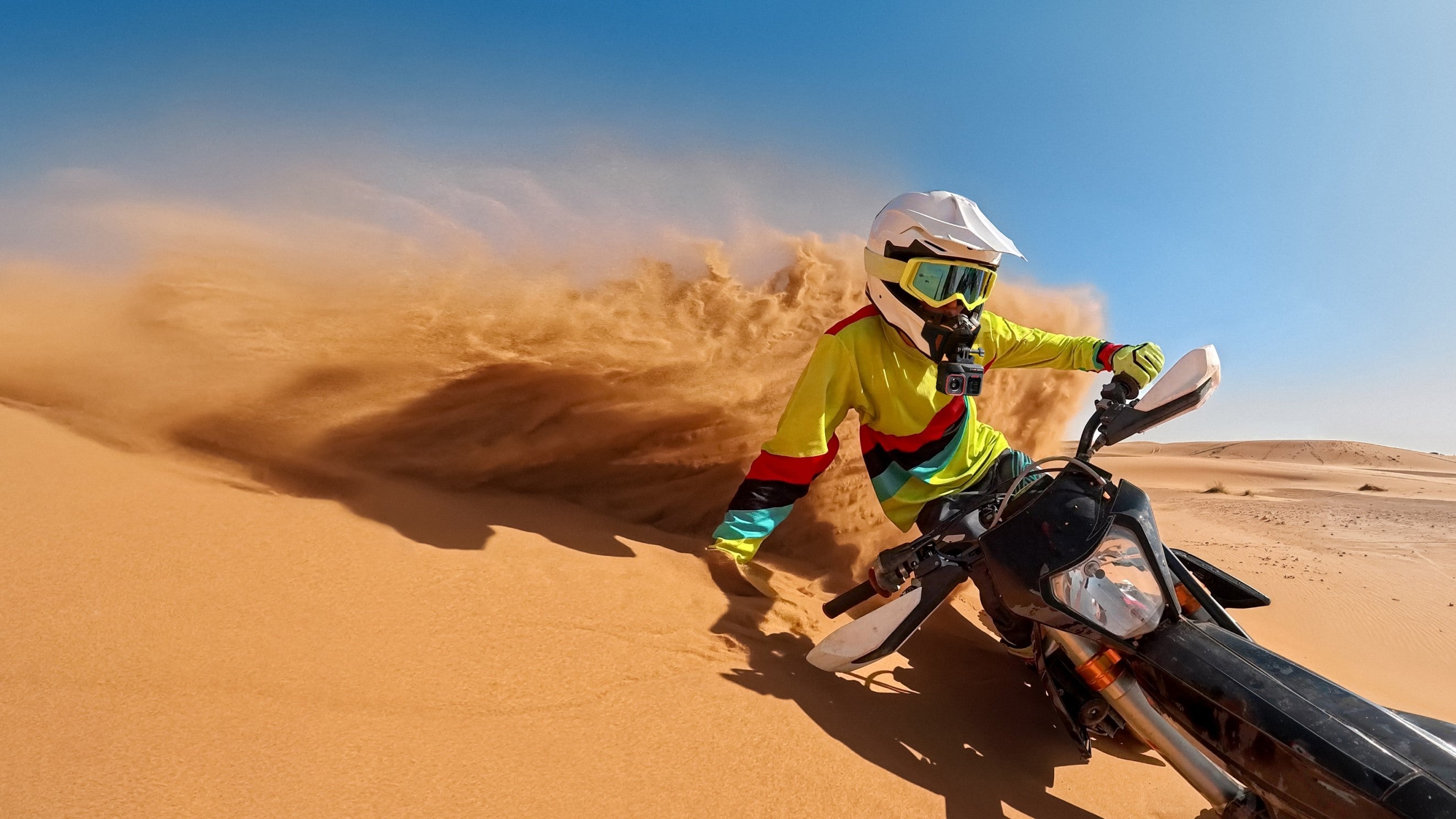 A person in motocross gear, including a white helmet and yellow jersey, is riding an off-road motorcycle in a sandy desert environment under a clear blue sky. The text 